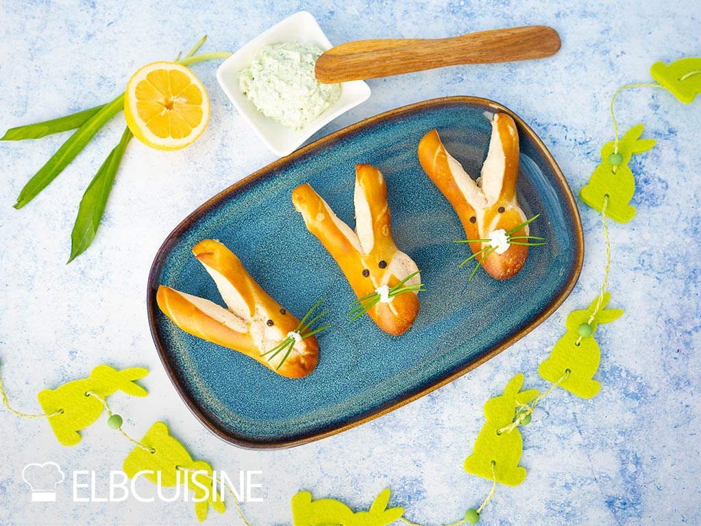 A plate of golden brown baked pretzel rolls in a cute bunny shape with long ears and a nose made of cream cheese and whiskers made of chives with a light green wild garlic dip ready for Easter brunch.